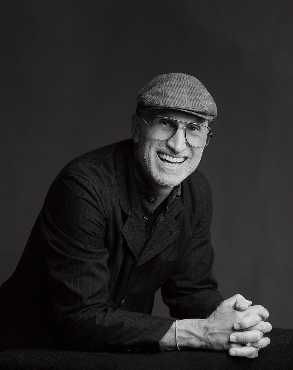 Smiling man in a flat cap and glasses, wearing a dark pinstripe suit jacket.
