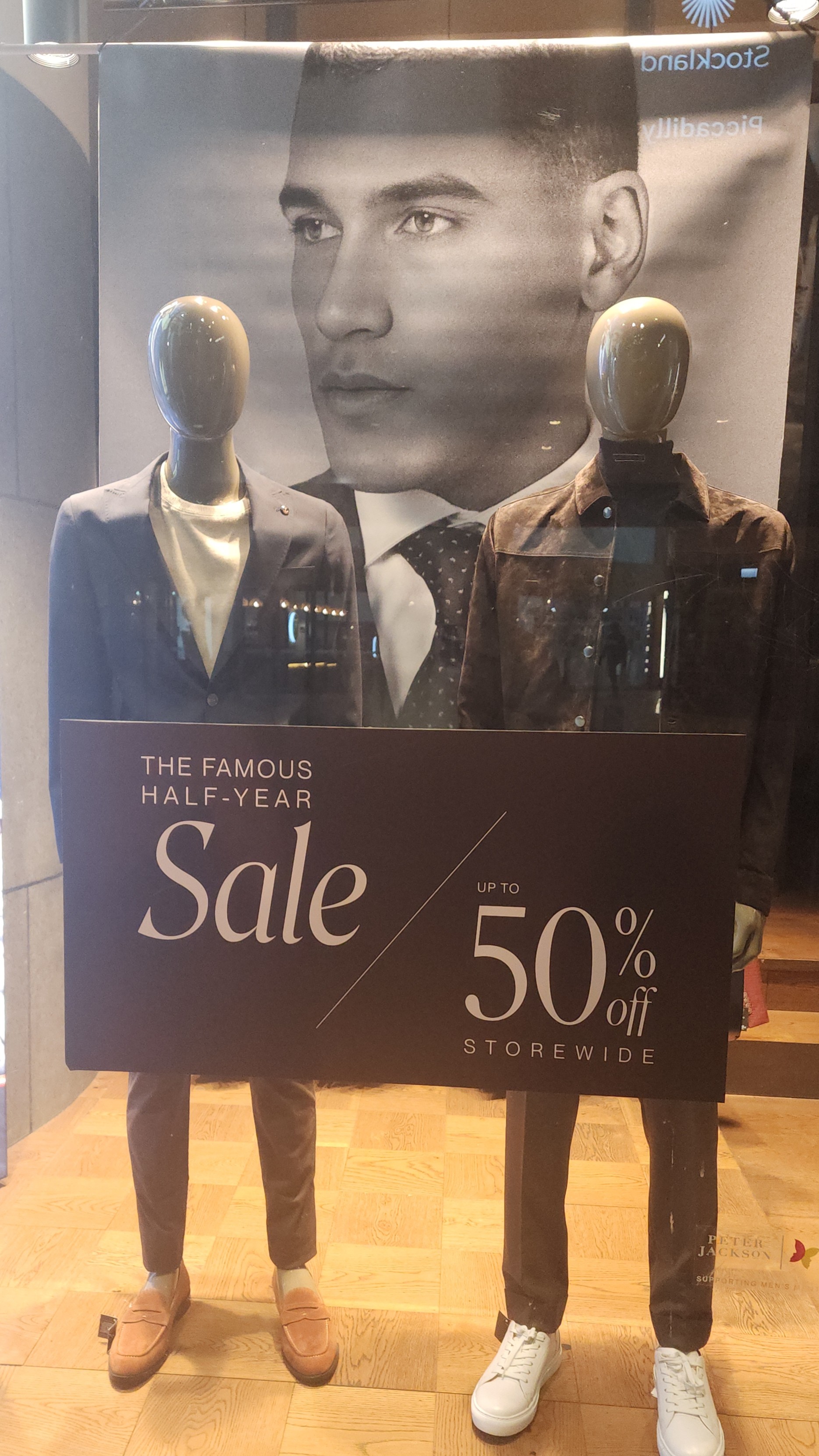 Two manneqins in suits and shoes and a large posters of a model in the background.