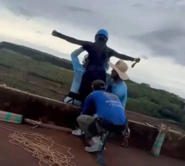 Three individuals are positioned on a rooftop, with one person standing and holding a long object, possibly a tool, while the ot
