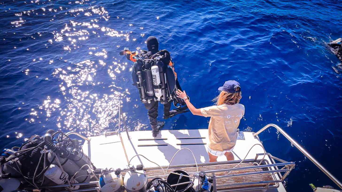 A scuba diver steps off a boat into the water as a nearby woman spots him