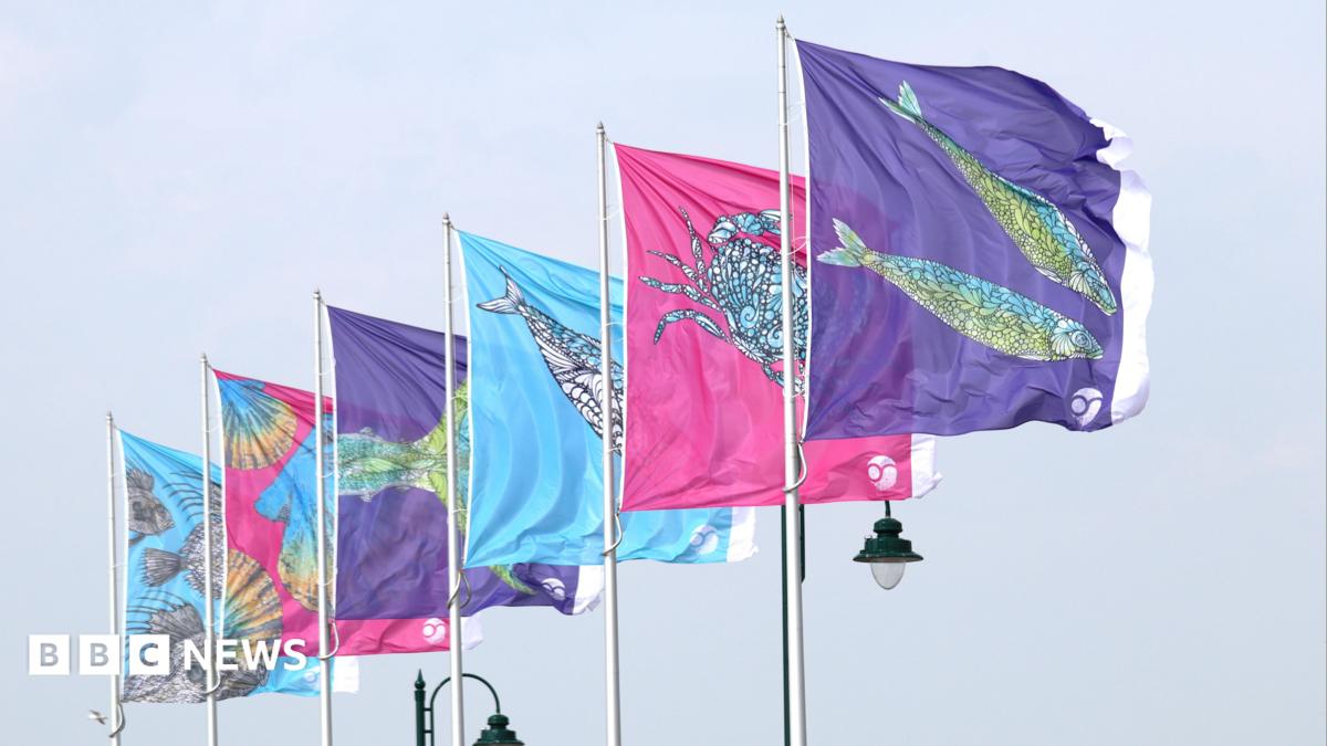 A row of colourful flags on tall poles, each decorated with stylised sea creatures, fly in the air against a pale sky.