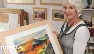 Woman with white hair holding a multi-coloured landscape painting showing hills and fields. She is wearing a grey dress with white top underneath