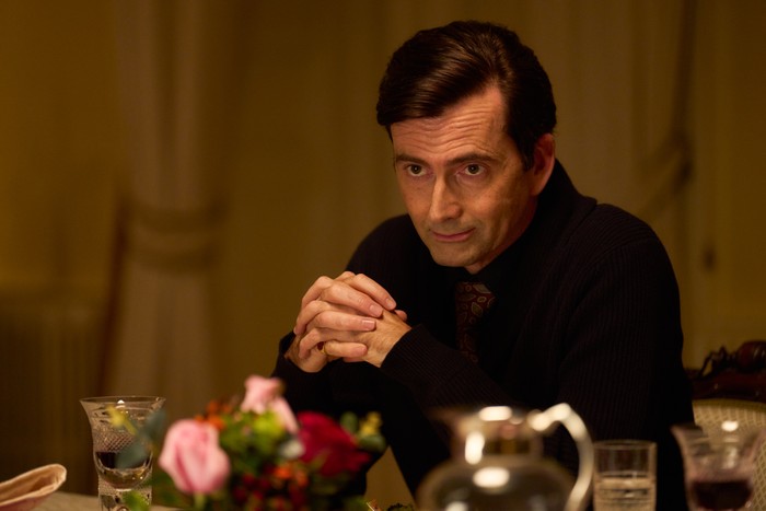 David Tennant in Rivals season 2 David Tennant in Rivals season 2, wearing a black shirt and red tie, sat at a dinner table.