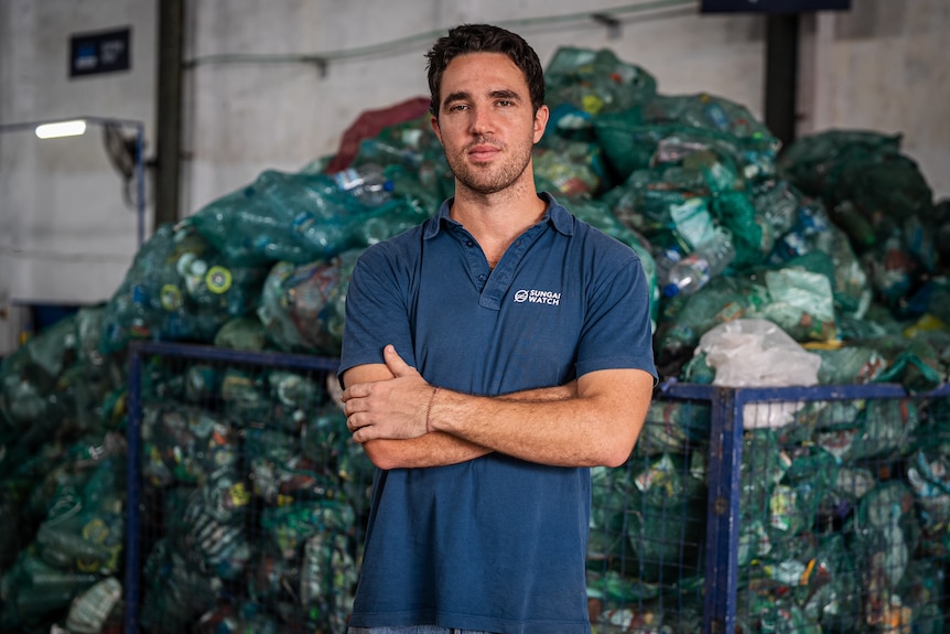 A close up of a man with his arms crossed wearing a blue polo shirt and standing infront of trash.