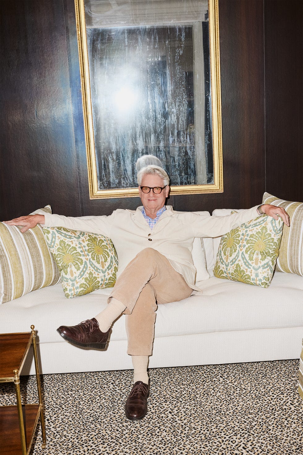 Man sitting on a couch with patterned pillows, hands outstretched, framed mirror behind him.