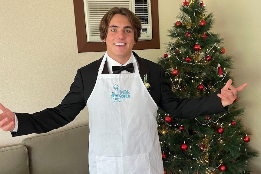 Luke wears an apron which reads "chef squid" and wears a suit infront of christmas tree.