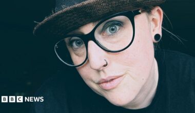 Kate Quinn wears a dark had a thick-rimmed glasses. She has blue eyes and a nose piercing and a large earring in one ear.