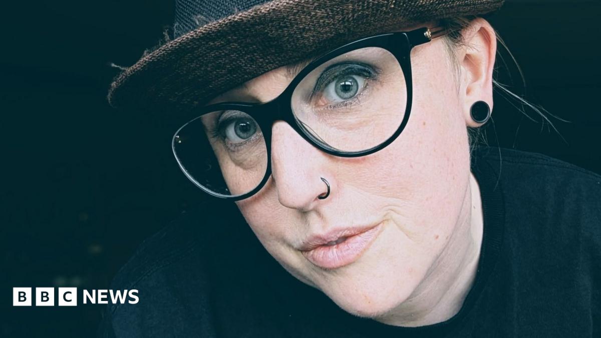 Kate Quinn wears a dark had a thick-rimmed glasses. She has blue eyes and a nose piercing and a large earring in one ear.
