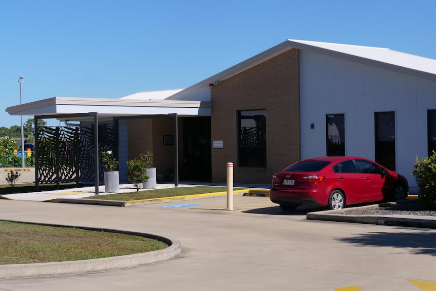 A small building with different coloured brick walls sits in front of a parking lot with a red car.