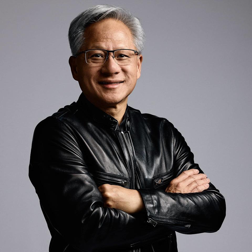 Jensen Huang stands with arms crossed, smiling, wearing a black leather jacket and thin-framed glasses against a gray studio background.