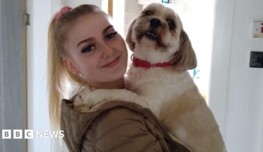 Blonde girl with high ponytail looking at the camera. She is wearing a green puffer jacket and holding a small white and tan dog. The dog is wearing a red collar.