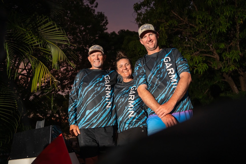 Three people (man, woman, man) standing together dark night, white light on them, all wearing blue / black longsleeve shirts