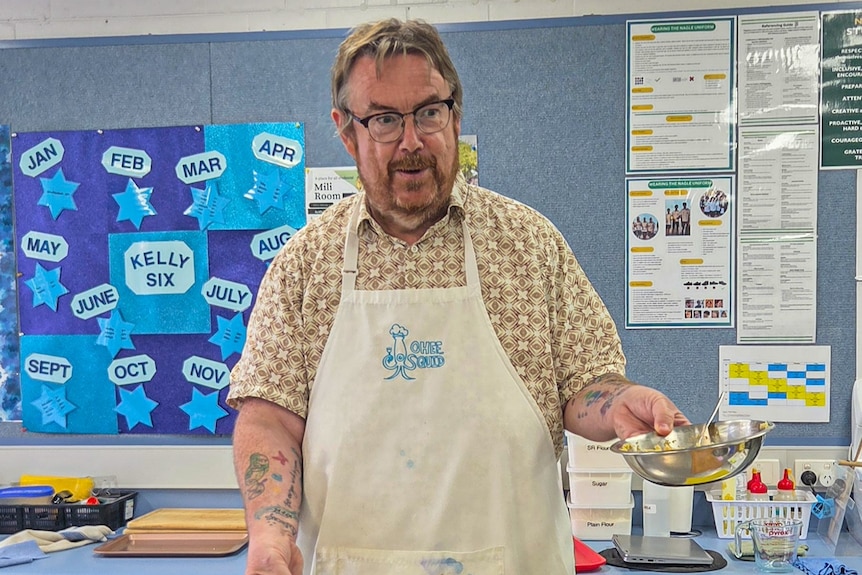 Derek Goforth wears an apron and holds a metal bowl in a class room. He has glasses and a beard, also a tattoo on right forearm.