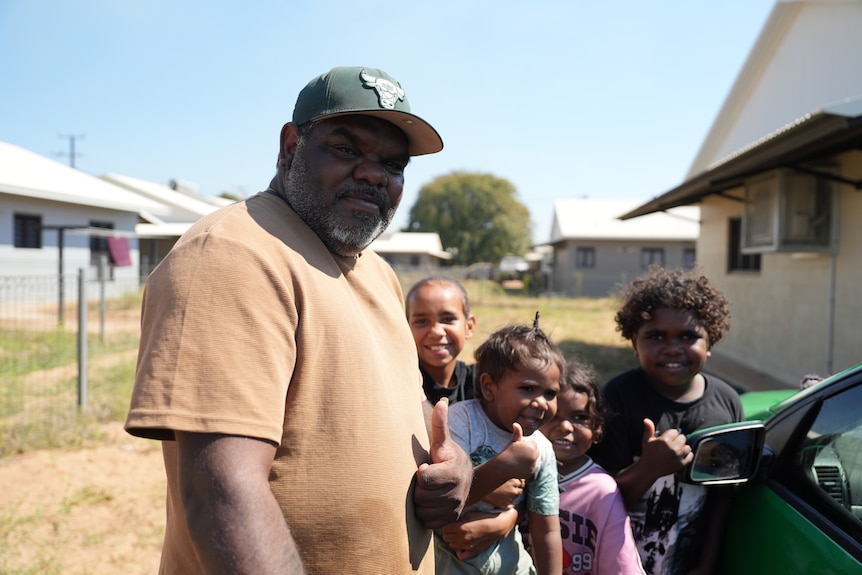 Aboriginal man, orange tshirt green cap, thumbs up standing next to four kids also smiling and thumbs up, houses behind them
