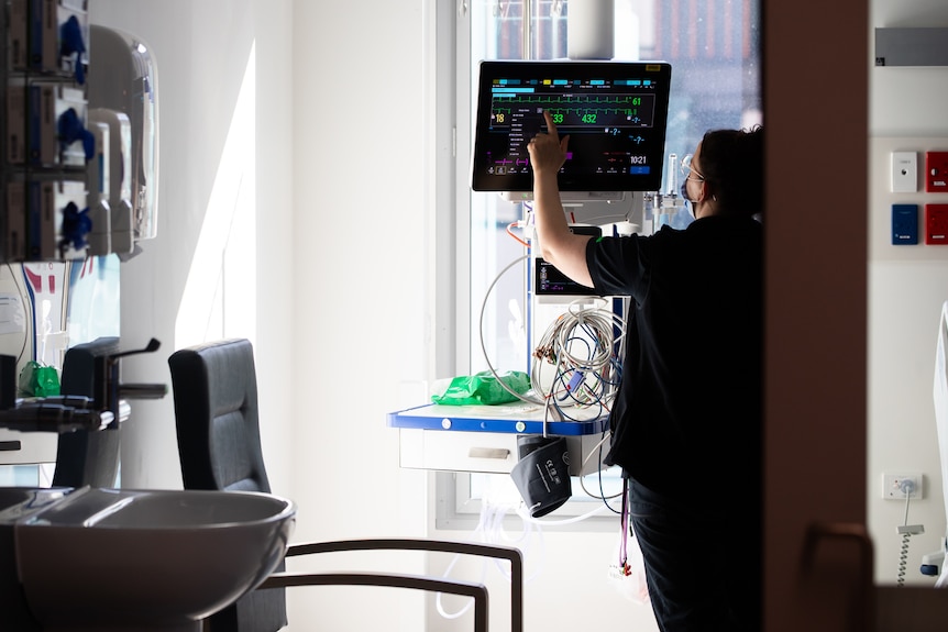 A nurse interacts with a monitor in a patient's room seen from outside.