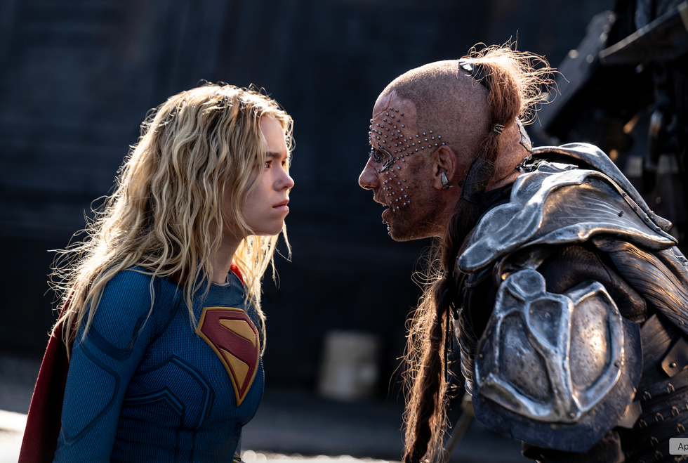 Superhero in blue and red costume confronts armored warrior with facial piercings.