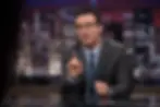 Man in suit and glasses speaks at a TV desk, raising one finger, with a blurred city skyline backdrop.