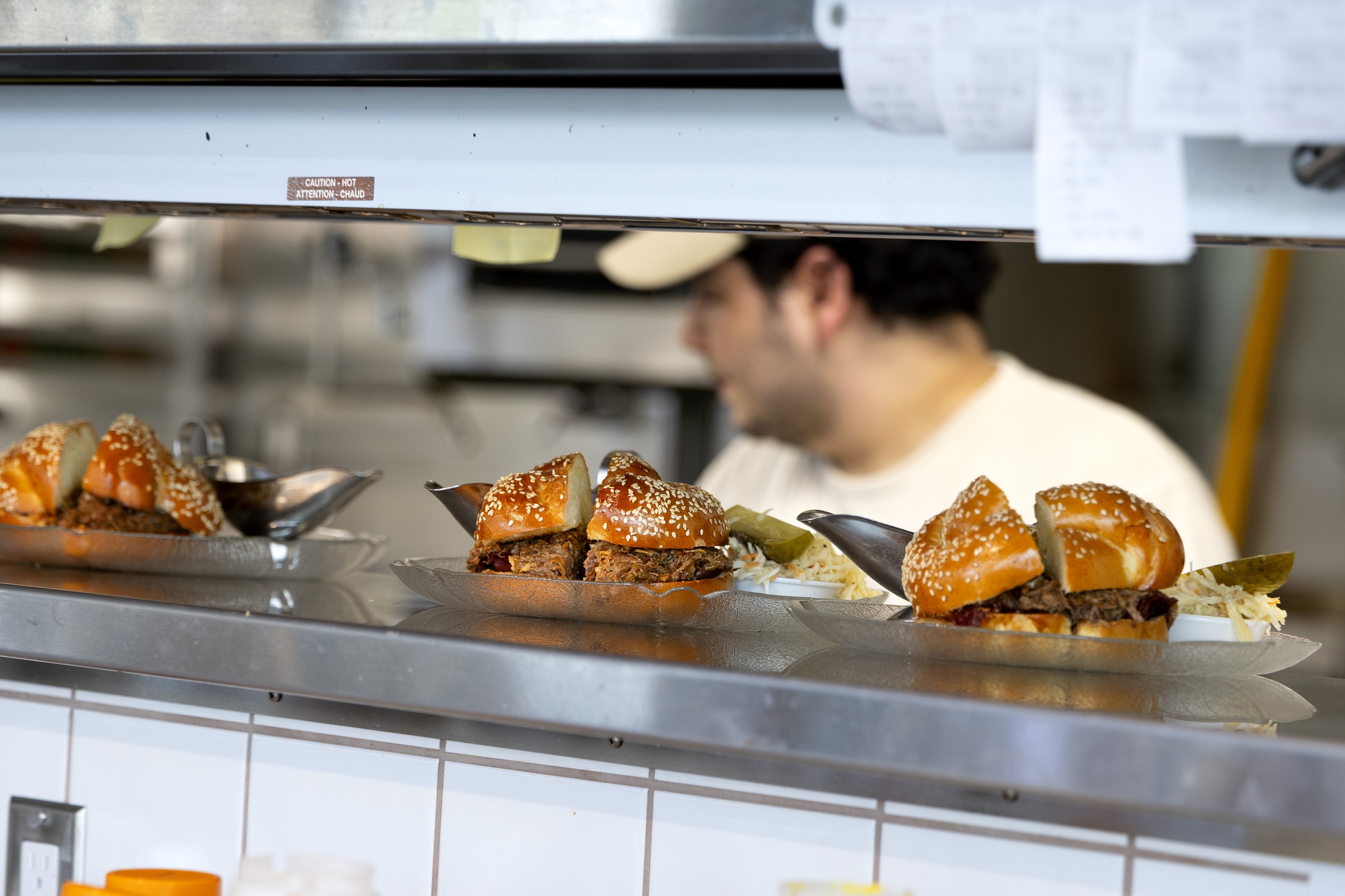 Three sandwiches under a heat lamp in a restaurant, with a kitchen worker in the background.
