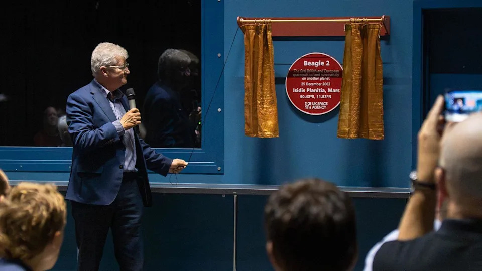 A red plaque is on a blue wall, revealed by a man with grey hair pulling on a rope that opened some gold curtains