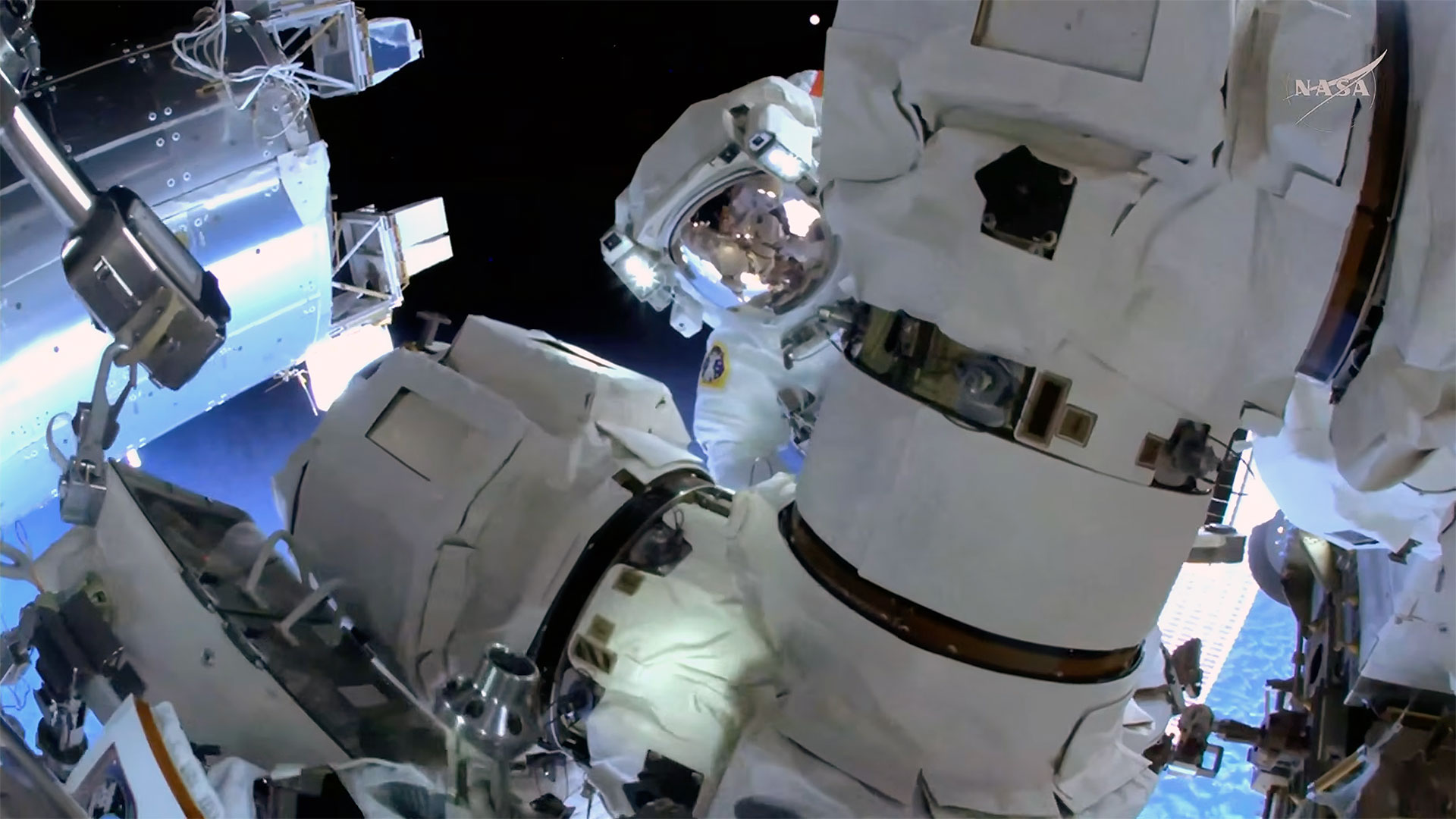 An astronaut in a white spacesuit peers around a robot arm outside of a space station