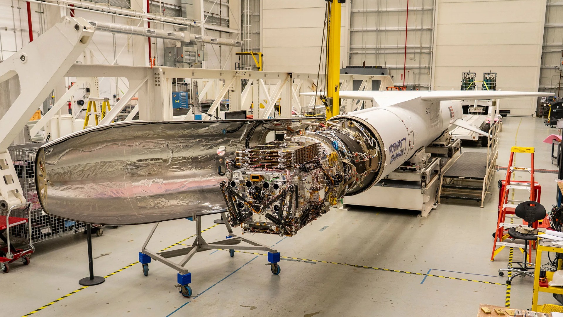 a long white rocket sits horizontal inside a hanger with its nose cone open, exposing a satellite with many components and actuators folded up into a dense rectangular shape