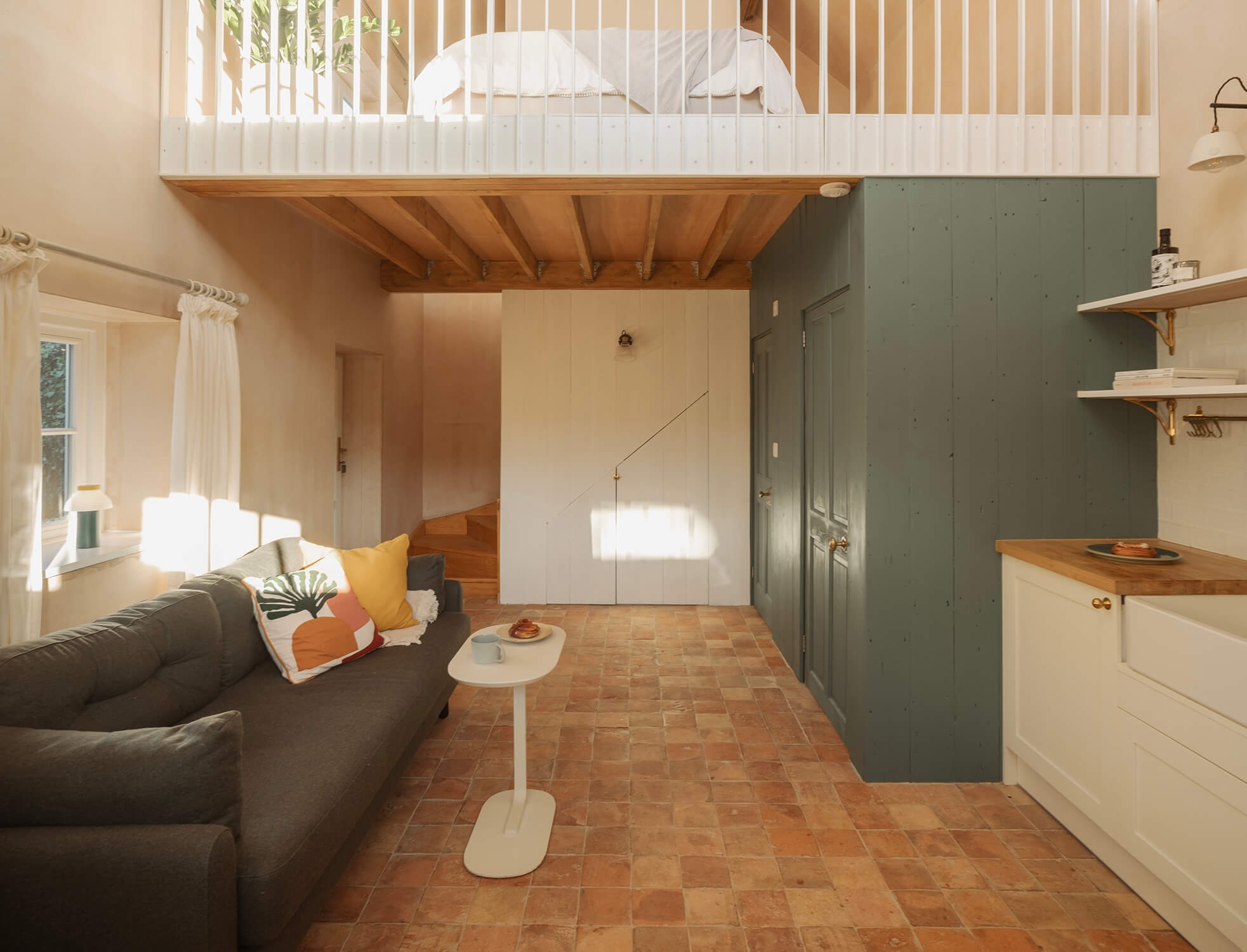 A contemporary split-level living space with brick floors, gray sofa, green storage joinery, white kitchenette, and a bedroom upstairs