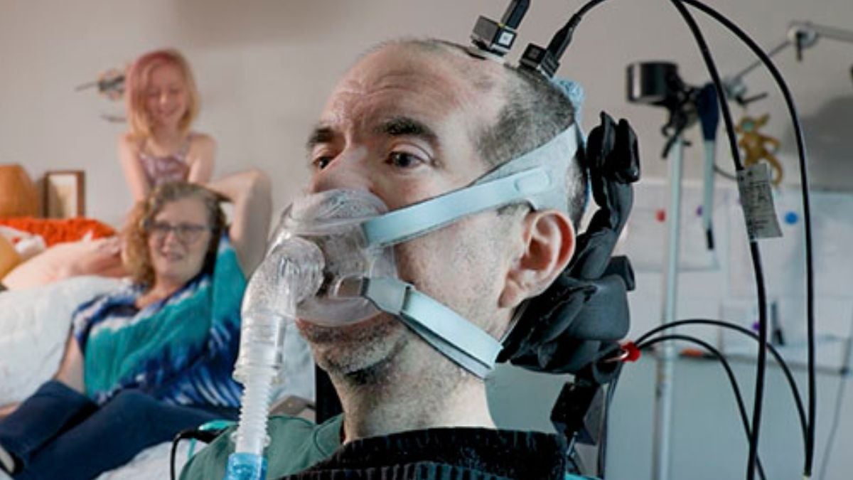 A man wearing a breathing mask and head-mounted brain-computer interface, with two family members blurred behind him.