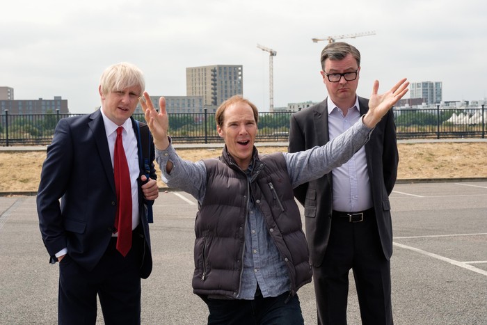 Richard Goulding as Boris Johnson, Benedict Cumberbatch as Dominic Cummings and Oliver Maltman as Michael Gove in Brexit: The Uncivil War, stood outside. Cummings is in the middle, with his arms outstretched, smiling.