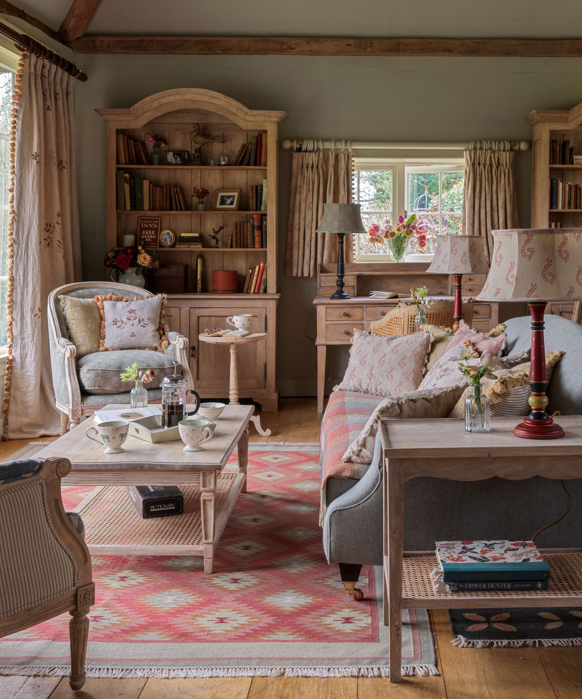 Cozy country living room with a gray sofa, geometric patterned rug, large wooden hutch, and a writing desk by a bright window.