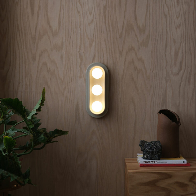 A modern wall sconce with three circular lights is mounted on a light wood paneled wall above a small table with books, a brown vase, and a plant.
