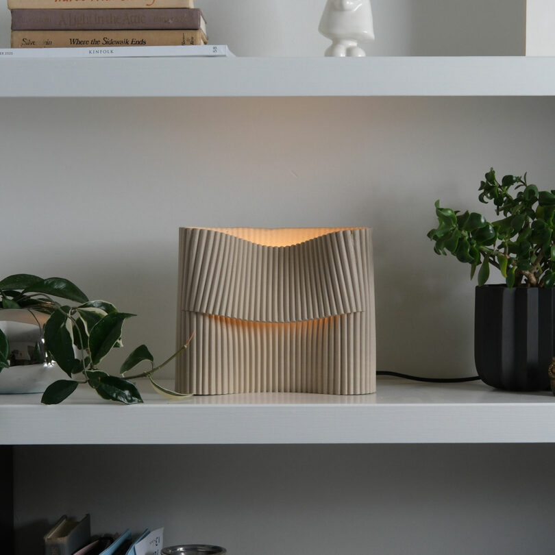 A beige, pleated Misewell table lamp sits on a white shelf, next to potted plants and books. The lamp emits a soft, warm light.