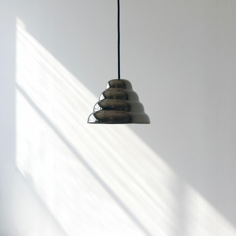 A dark, dome-shaped pendant light hangs from the ceiling against a plain white wall, with sunlight casting diagonal shadows across the surface.