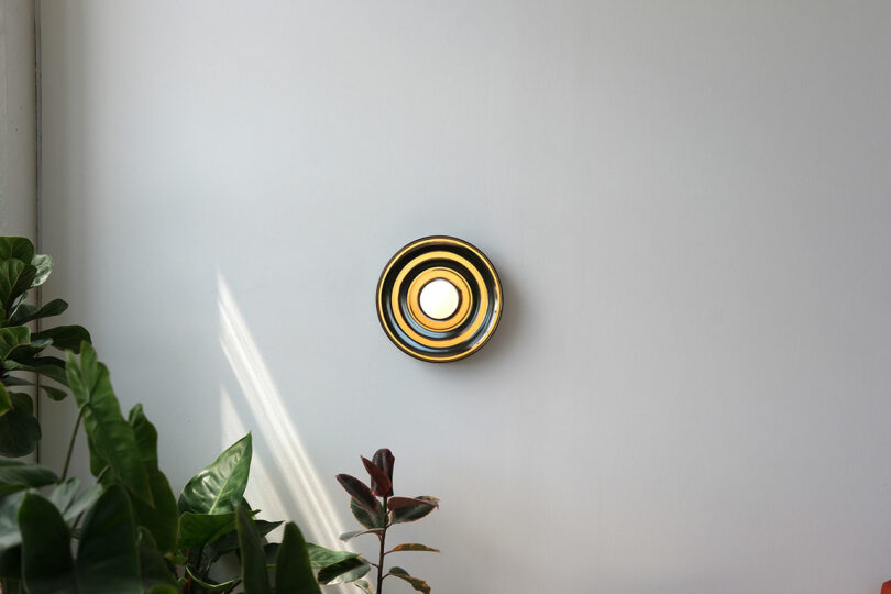 A round, modern wall light with concentric rings emits a warm glow on a plain white wall, with several green and red potted plants nearby.