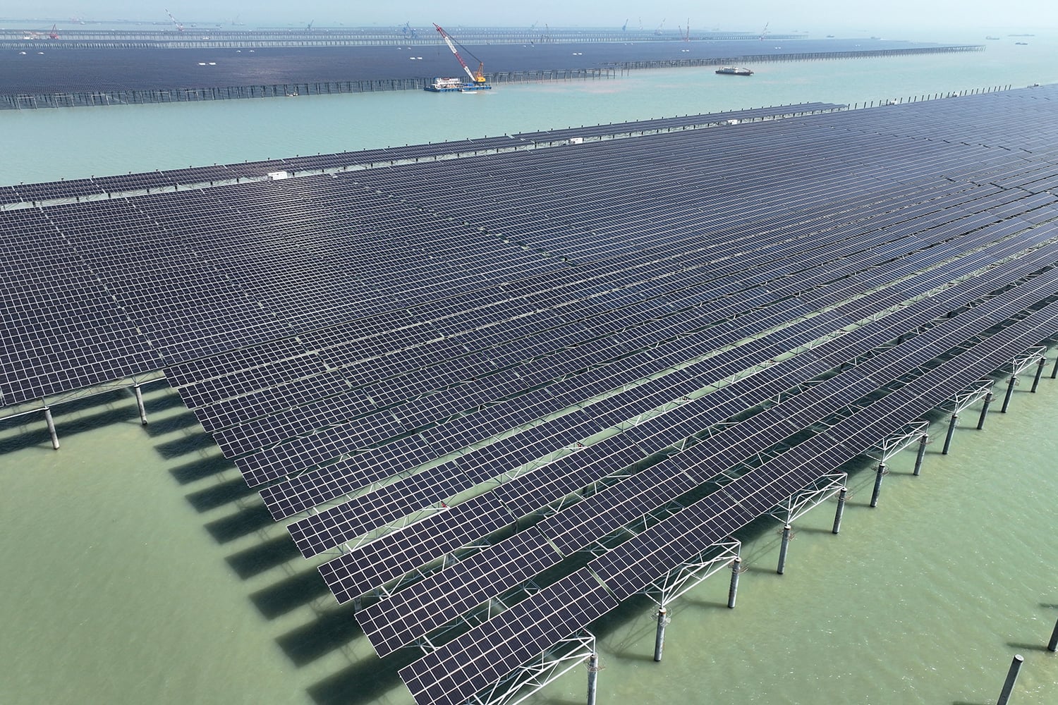 Rows of solar panels sit in a tidal pool during the daylight. A crane is in the background