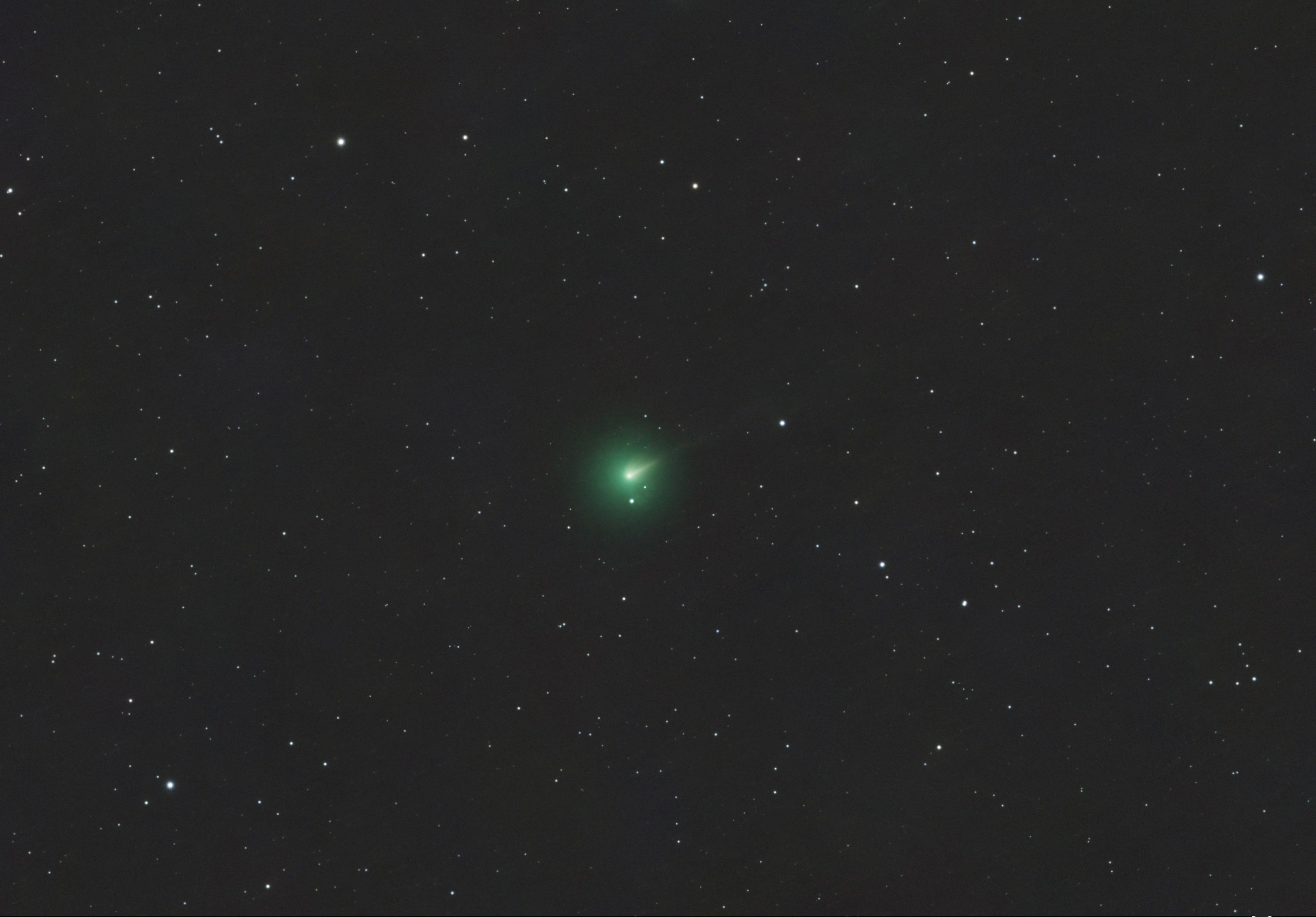 A telescope image of Comet 220P in outburst, showing a bright green coma glowing near the center of the frame against a dark star-filled background. The comet's nucleus appears as a bright condensed point within the fuzzy green cloud, with a faint tail extending to the upper right.