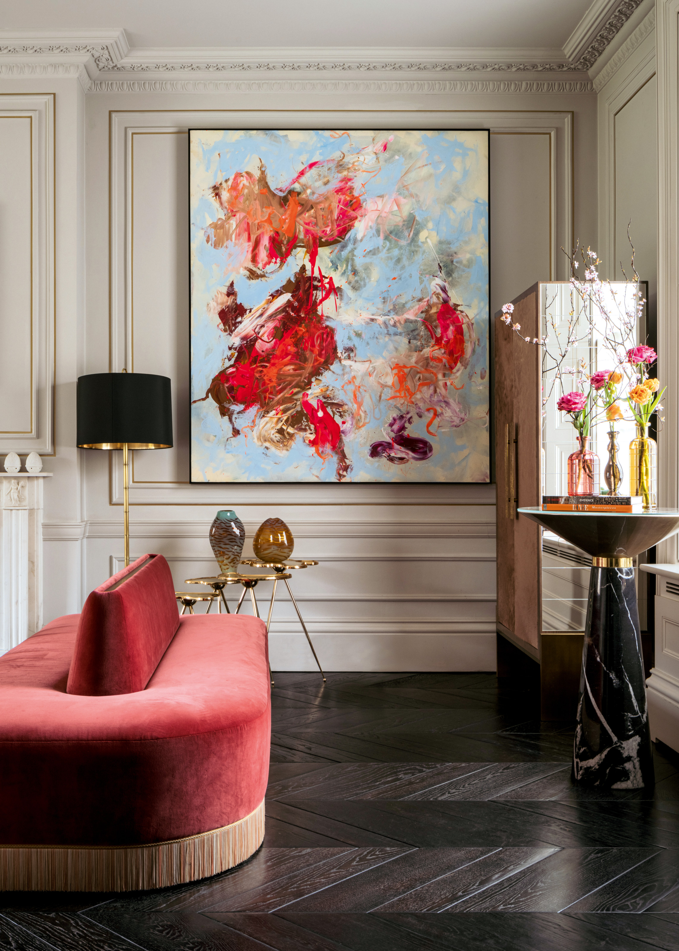 Corner of a room with a marble bar cabinet, black marble cocktail table, dark wood chevron flooring, red velvet seating and abstract artwork