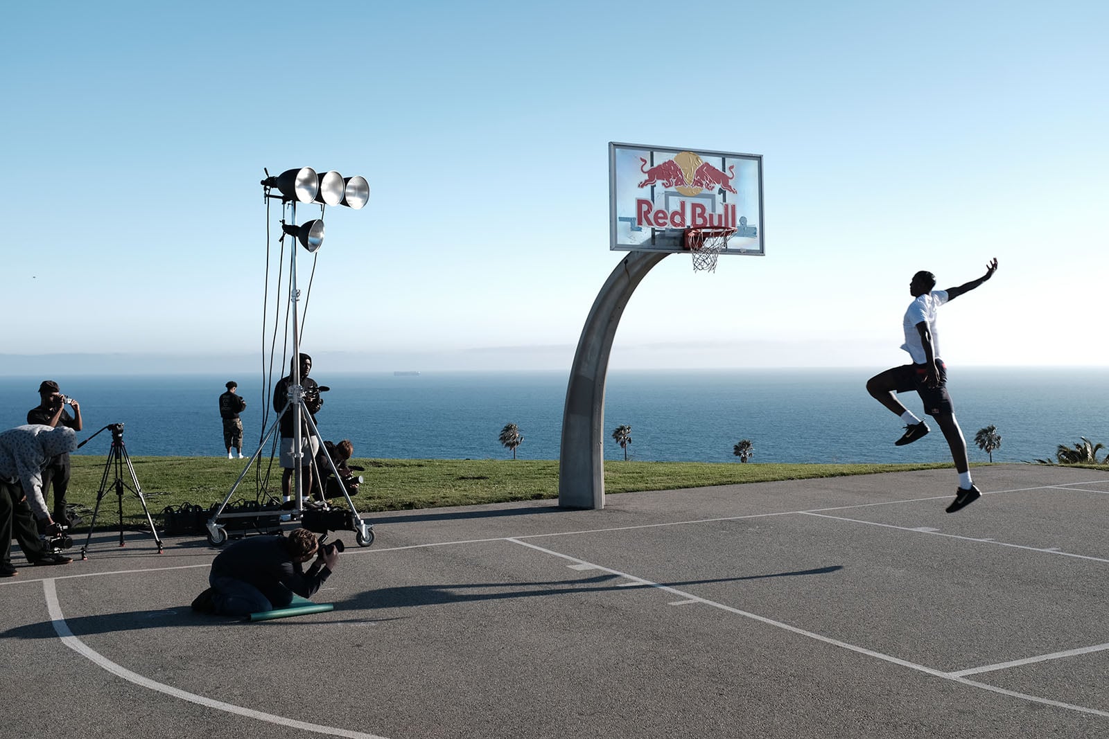 A basketball player leaps toward a Red Bull-branded hoop on an outdoor court by the ocean, while photographers and camera crews capture the moment under bright lights.