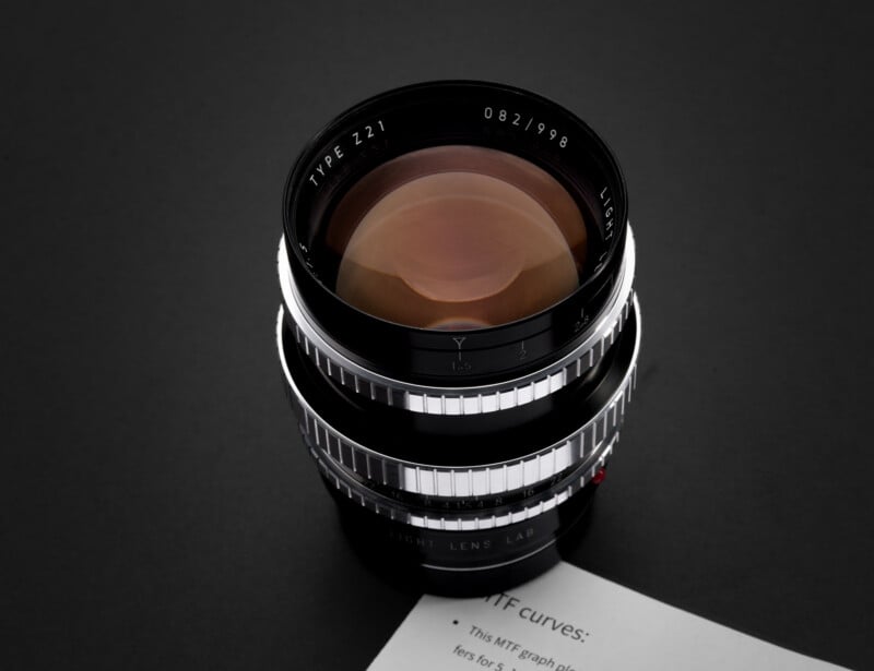 A camera lens with silver and black detailing is placed on a dark surface. Part of a white paper with partial text is visible in the lower right corner of the image.