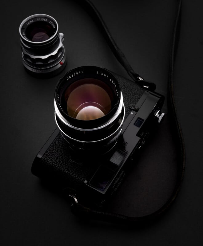 A black vintage camera with a large lens and a leather strap lies on a dark surface, next to an additional detachable camera lens.