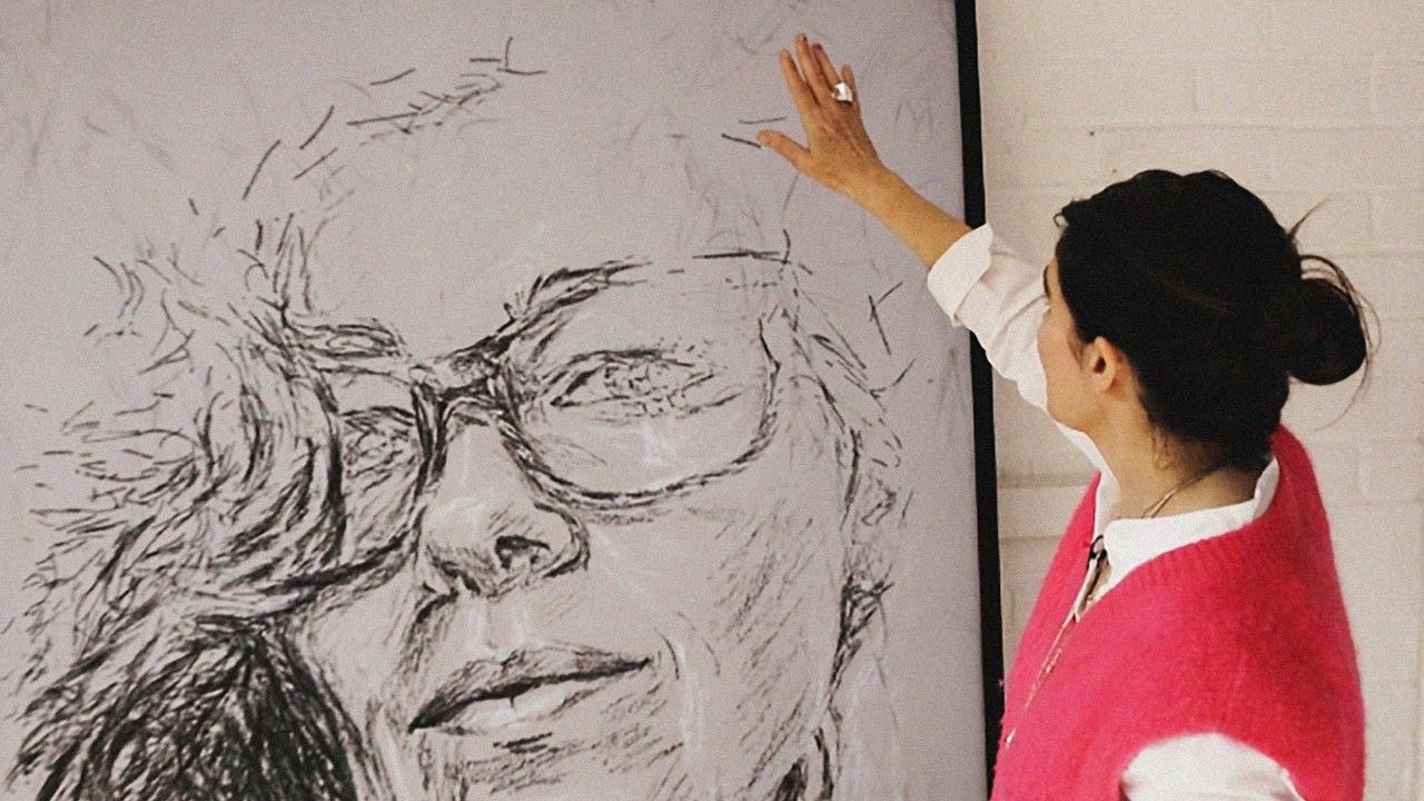 A woman with dark hair tied back, wearing a pink vest over a white shirt, reaches up to touch a large, expressive sketch of a face on a canvas.