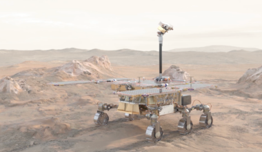 Boxy rover up on 6 metallic wheels in a rocky, dusty landscape. It has wide solar panels.