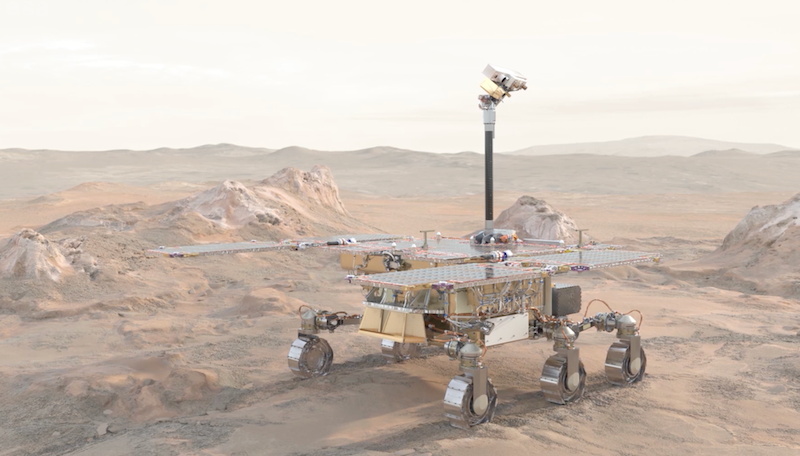 Boxy rover up on 6 metallic wheels in a rocky, dusty landscape. It has wide solar panels.