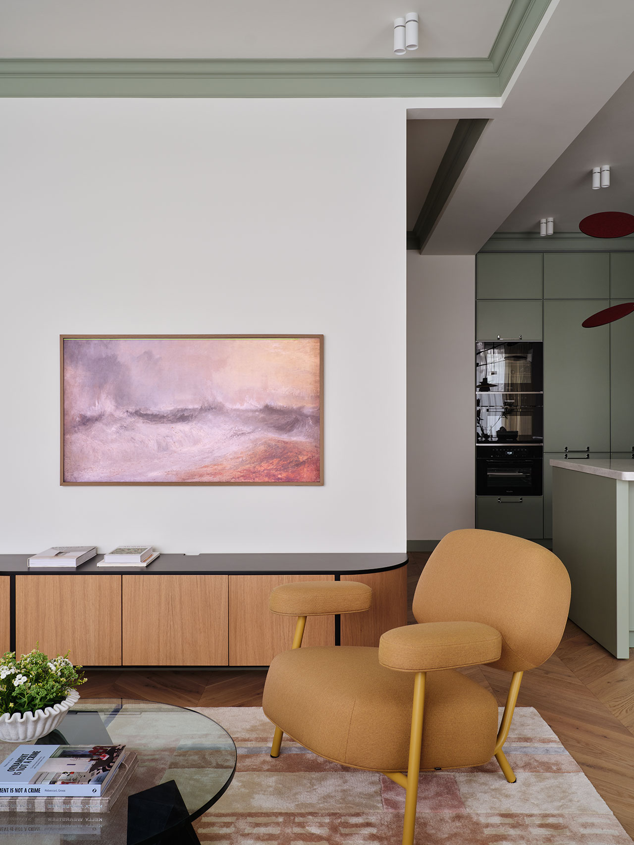 A modern living room with a mustard armchair, abstract wall art, a wood sideboard, and a view into a kitchen with green cabinets.