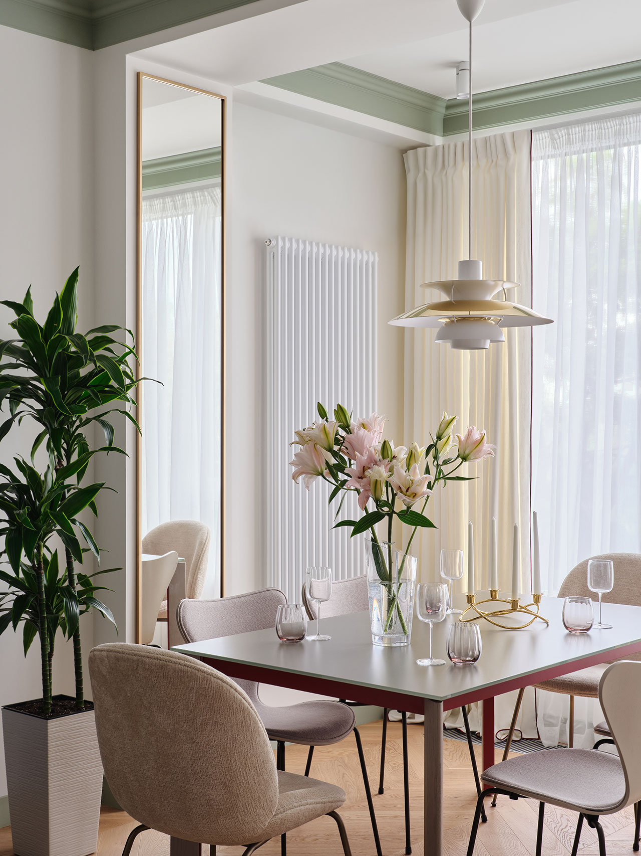 Modern dining room with a rectangular table, beige chairs, a vase of pink lilies, a tall mirror, a potted plant, and large windows with sheer curtains.