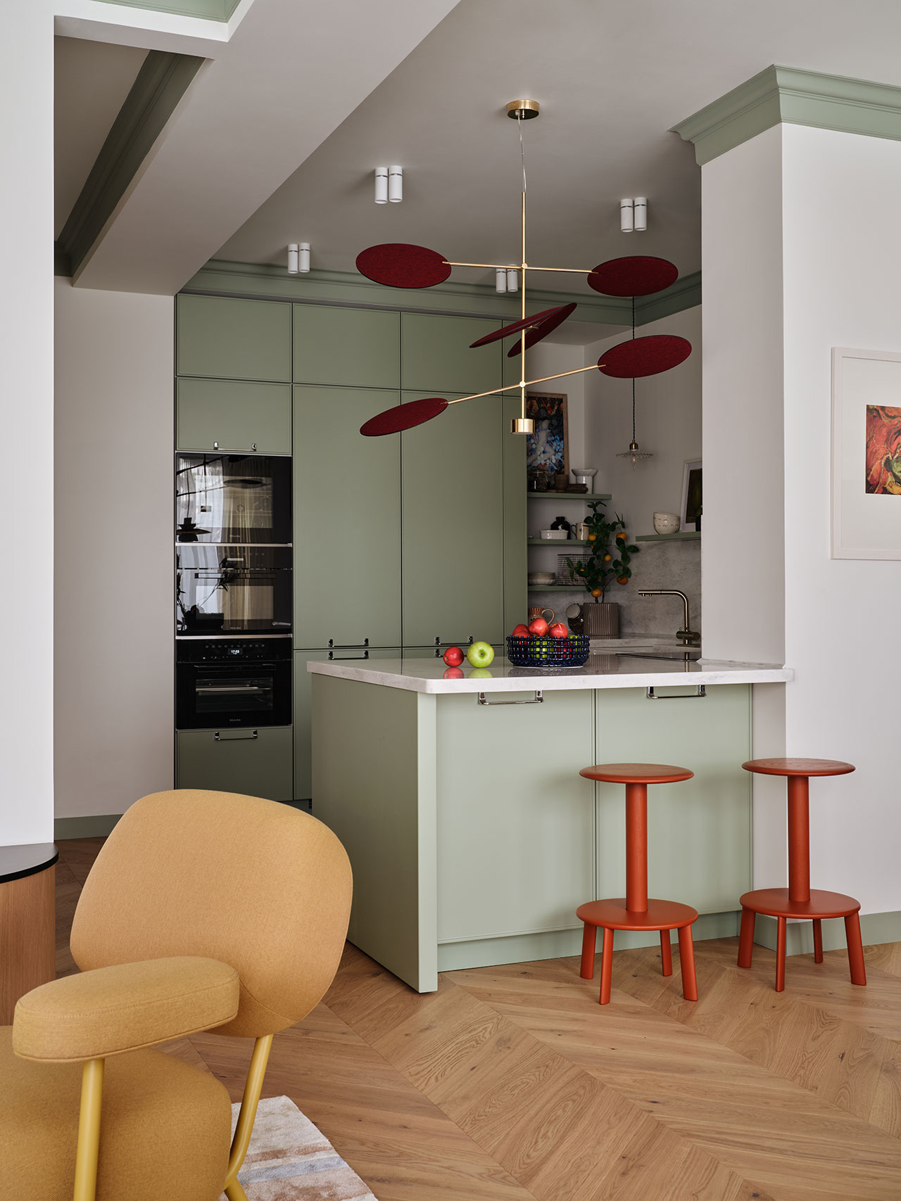 Modern kitchen with sage green cabinets, a white countertop island, two red stools, contemporary pendant light, and a mustard yellow chair on herringbone wood flooring.
