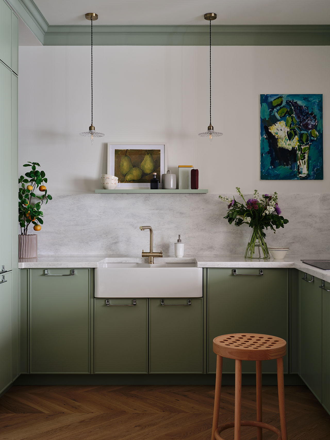 Modern kitchen with sage green cabinets, white farmhouse sink, marble backsplash, wooden stool, potted plant, and wall art on white walls.