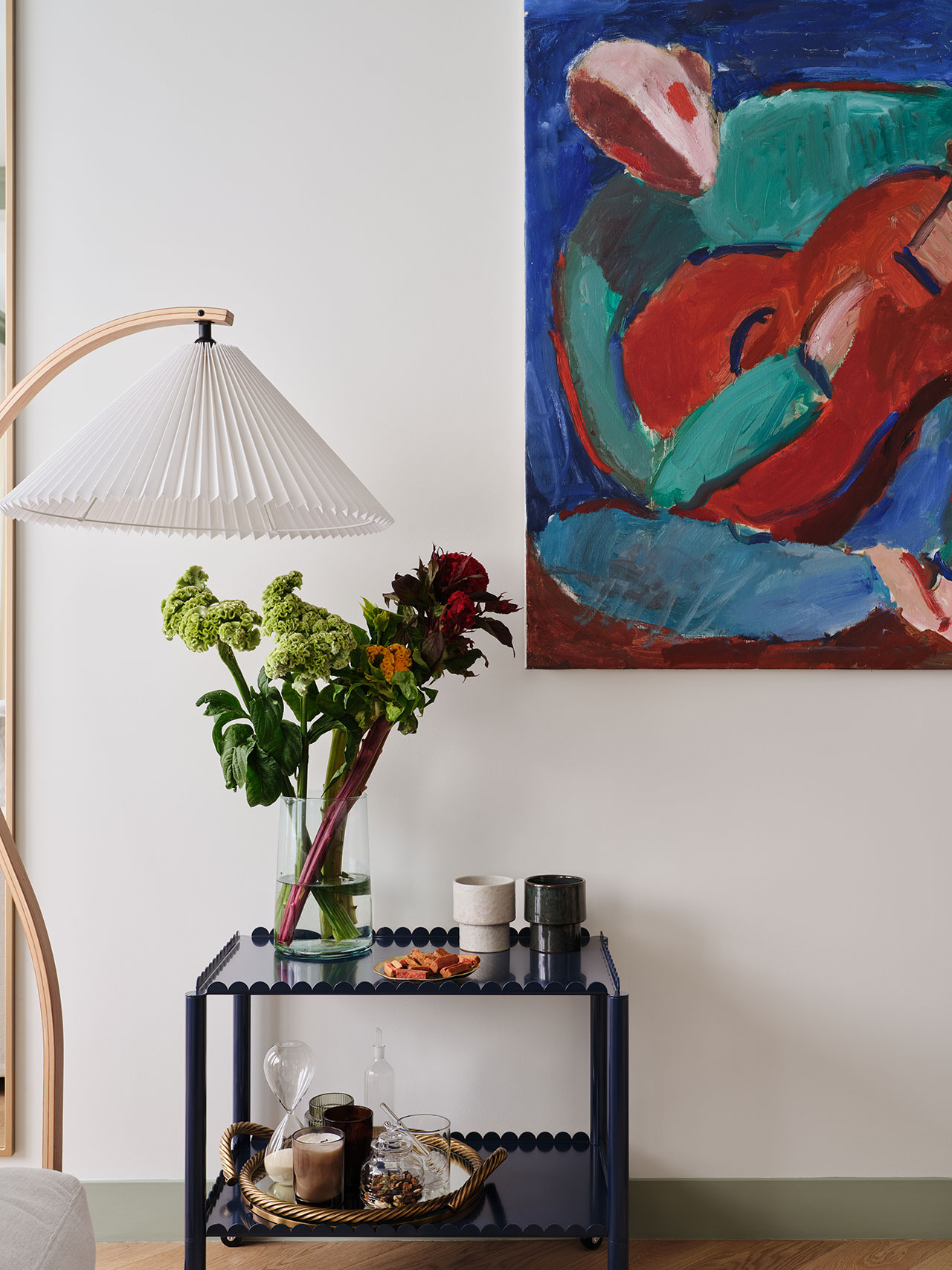 A floor lamp, a vase with flowers, and a small cart with various items sit below a colorful abstract painting on a white wall.