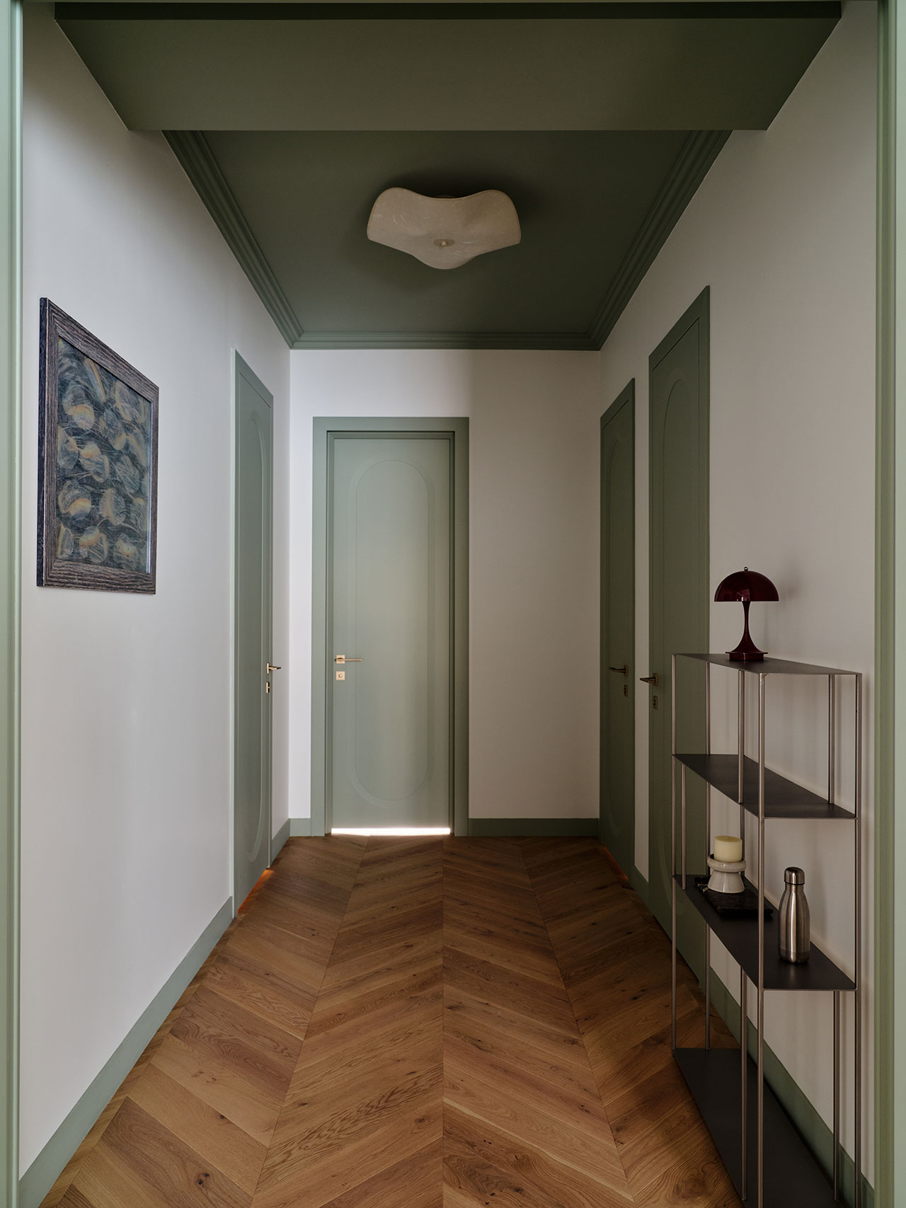 A hallway with green doors and trim, wood herringbone flooring, a ceiling light, a framed picture on the wall, and a metal shelf holding decor items.