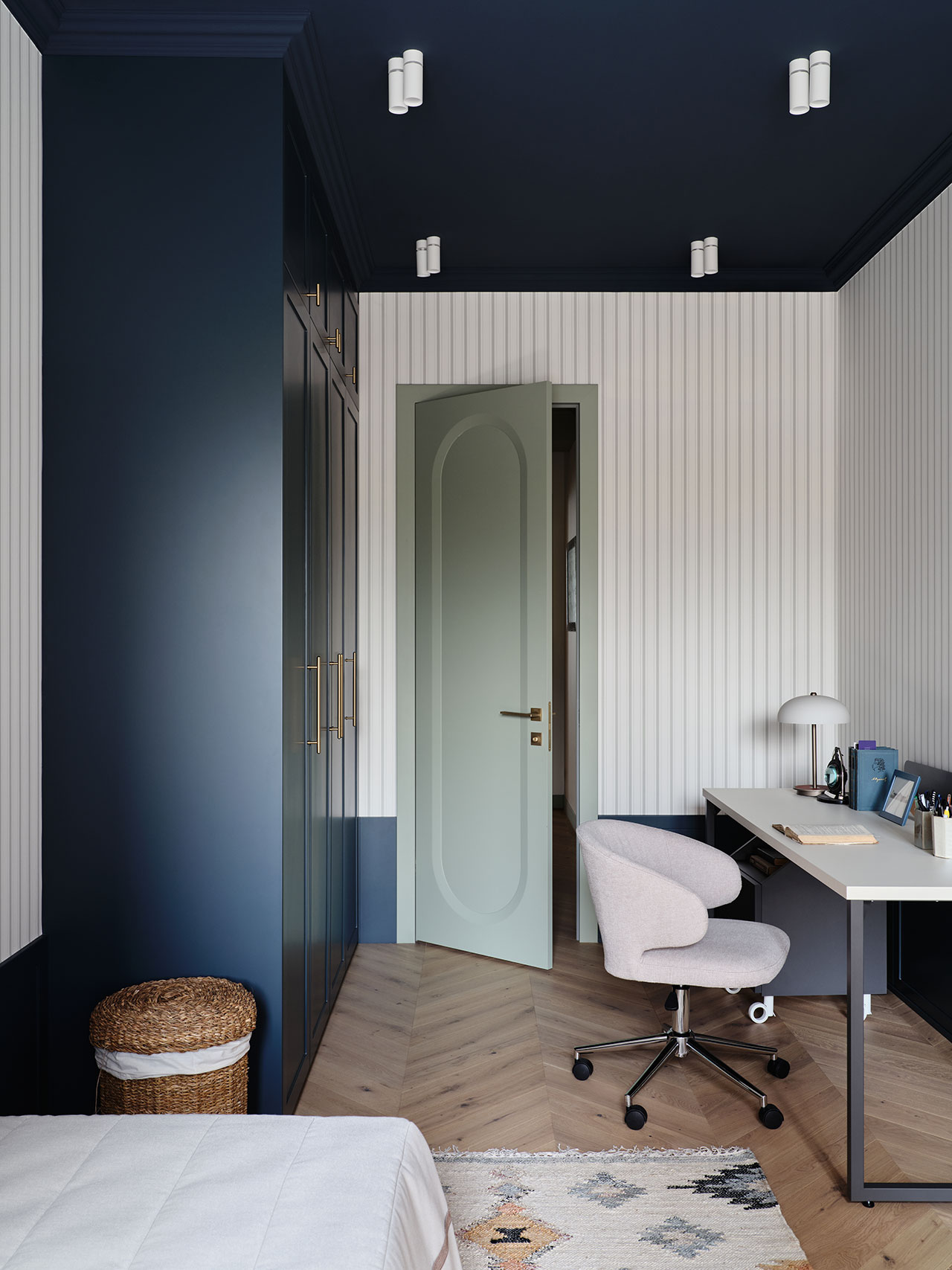 Modern home office with a desk, swivel chair, and lamp, featuring dark blue walls, light wood floors, and a partially open door. A basket and closed wardrobe are also visible.