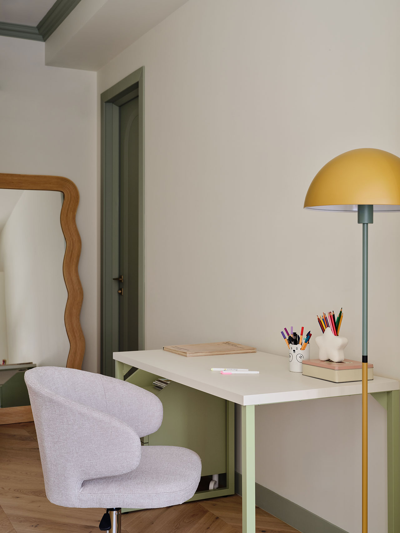 A modern desk with art supplies, a sculptural mug, a notepad, and a yellow lamp sits beside a light gray chair and a wavy-framed mirror in a minimalist room.